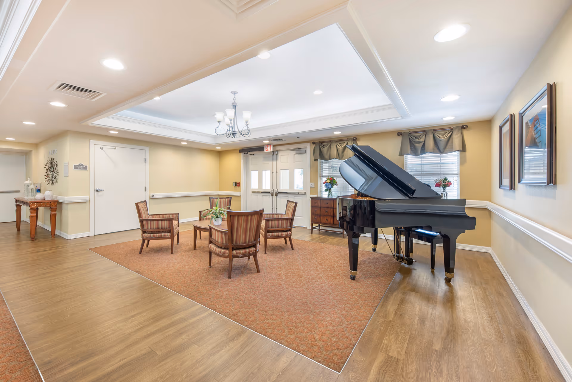 A bright and spacious common area in a senior living facility featuring a black grand piano, a round wooden table with four striped upholstered chairs, a wooden sideboard with flower arrangements, and framed artwork on the walls. The room has wood flooring with a large area rug, recessed lighting, and a chandelier in a tray ceiling. Double doors and windows with valances are visible in the background.