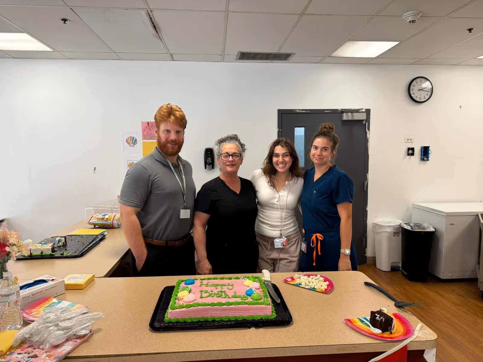 Four people standing behind a counter in a room, smiling at the camera. On the counter in front of them is a birthday cake decorated with green and pink icing, a plate with popcorn, and a plate with a piece of chocolate cake. The room has a white wall, a clock, and a door in the background.