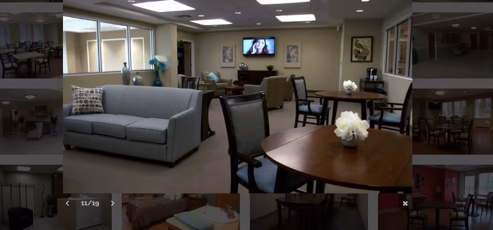 A senior living facility common area with a gray sofa, several wooden tables with chairs, and a wall-mounted TV. The room is well-lit with ceiling lights and has decorative vases and framed artwork on the walls.