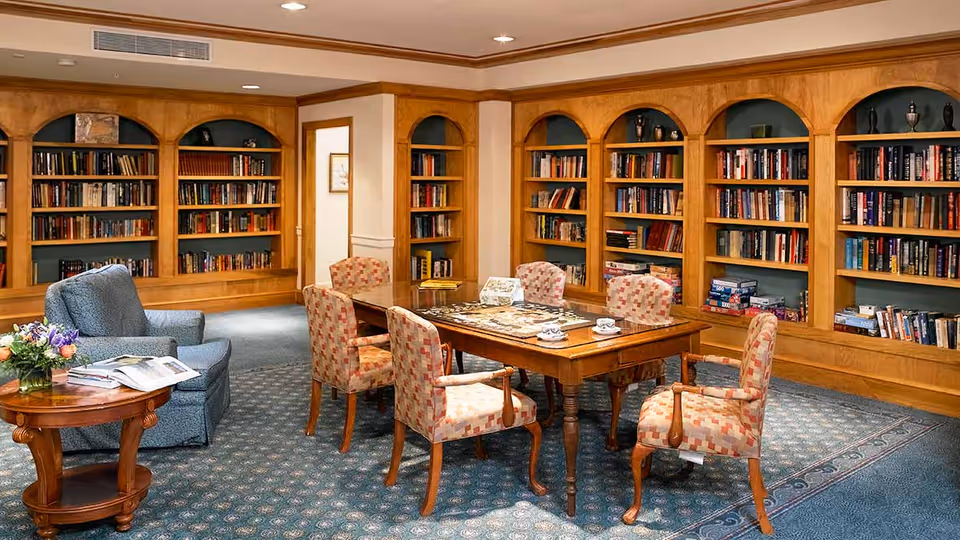 A cozy library or reading room with wooden bookshelves filled with books and board games lining the walls. In the center, there is a wooden table with six upholstered chairs arranged around it, and a puzzle or board game set up on the table. To the left, there is a blue armchair next to a small round wooden side table with a flower arrangement and some open books. The room has a patterned carpet and warm lighting.