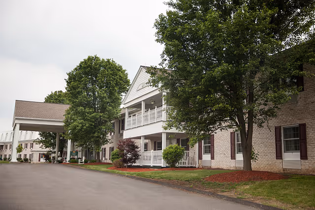 Exterior view of a senior living facility building with a covered entrance, multiple windows with shutters, balconies, and landscaped greenery including trees and bushes along the driveway.