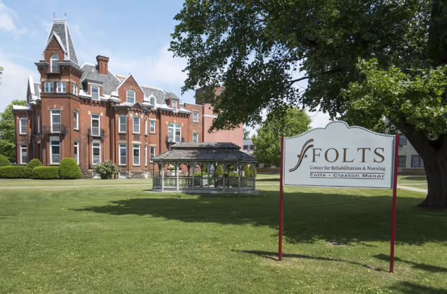 A large historic red brick building with multiple windows and a steep roof, situated on a well-maintained lawn with a wooden gazebo in the middle. A sign in the foreground reads 'Folts Center for Rehabilitation & Nursing Folts - Claxton Manor'. Trees provide shade on the right side of the image under a partly cloudy sky.
