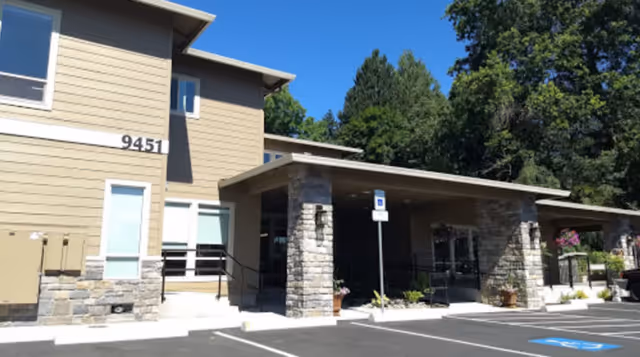 Exterior view of a modern residential care facility building with beige siding and stone pillars supporting a covered entrance. There is a parking lot with marked spaces, including a handicapped parking spot, and trees in the background.