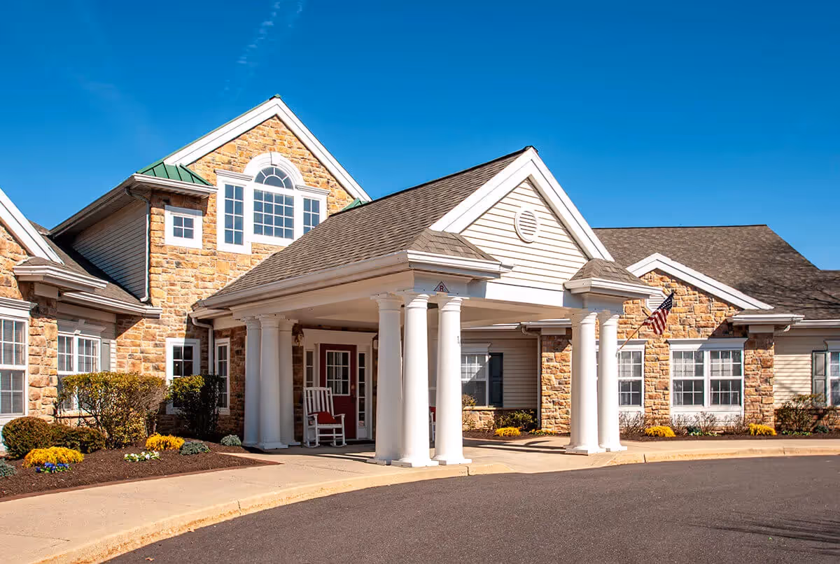 Exterior view of a senior living facility with stone and siding walls, white columns supporting a covered entrance, rocking chairs on the porch, and an American flag mounted near the windows under a clear blue sky.