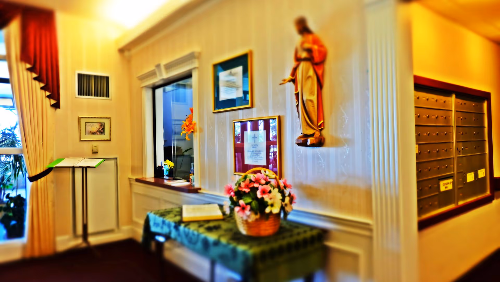 Interior view of a reception or lobby area in a retirement community featuring a small table with a basket of flowers, framed pictures and notices on the wall, a statue mounted on the wall, a window with a counter, and a set of mailboxes on the right side.