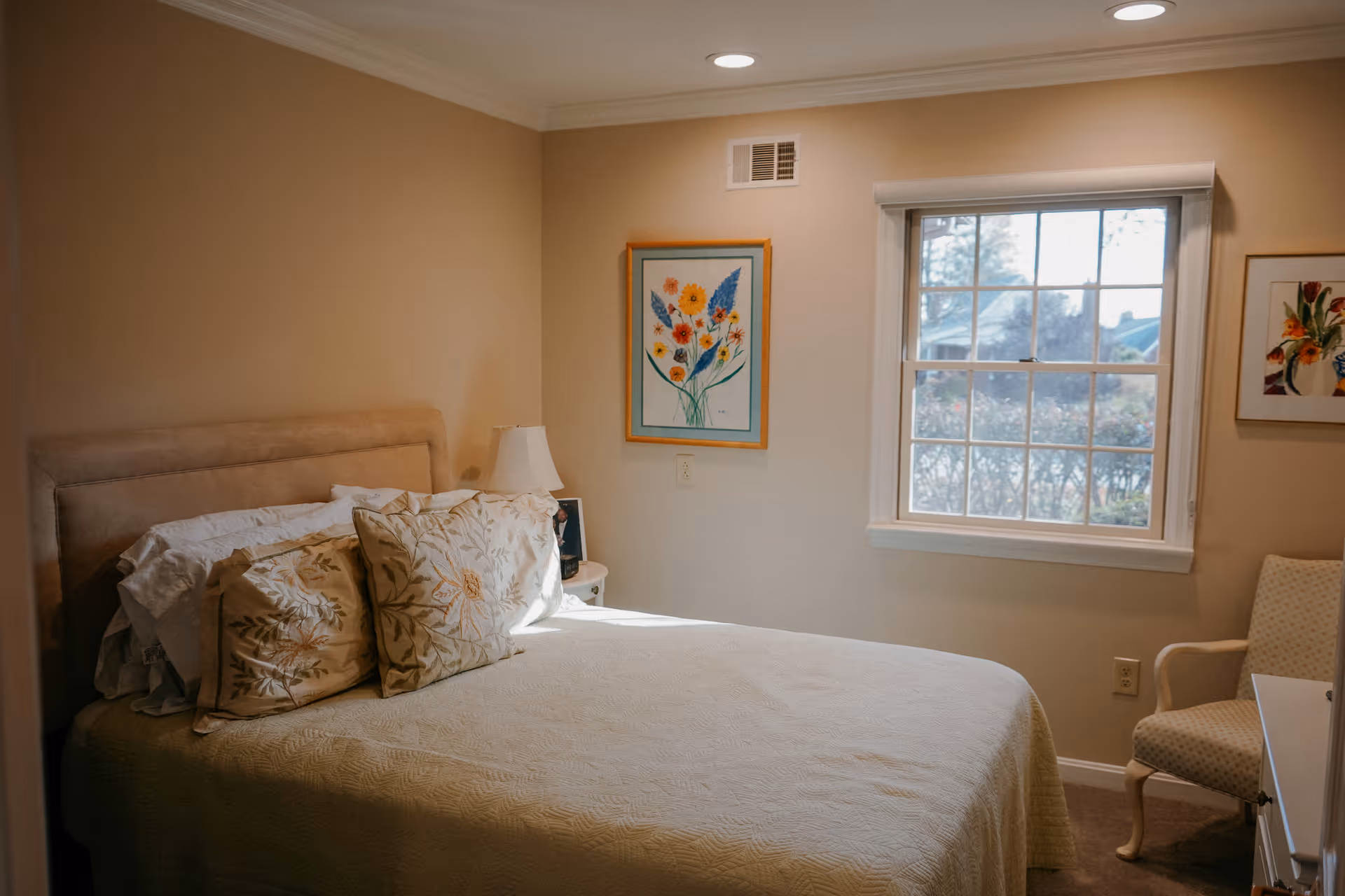 A cozy bedroom with a bed covered in a light-colored quilt and floral pillows. There is a bedside table with a lamp and a framed photo. The room has beige walls, a window with a view of bushes and trees outside, and two framed floral paintings on the walls. A cushioned chair is positioned near the window.