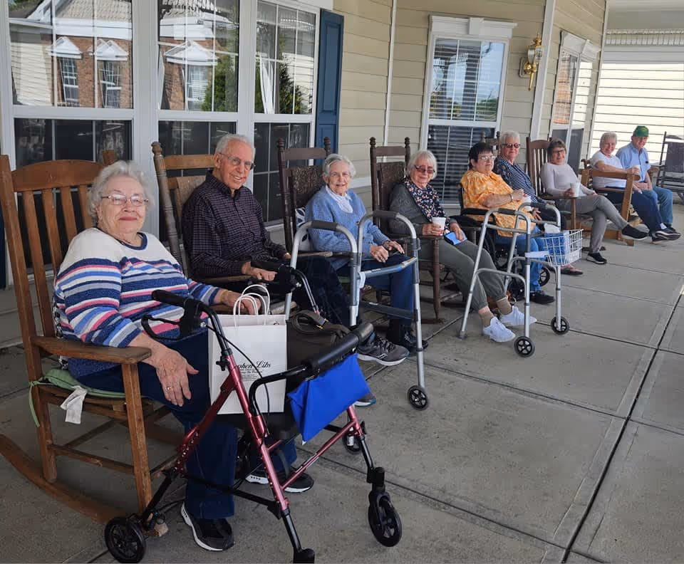 A group of elderly people sitting on rocking chairs on a covered porch outside a building. Some have walkers or mobility aids with them, and they appear to be enjoying a social moment together.