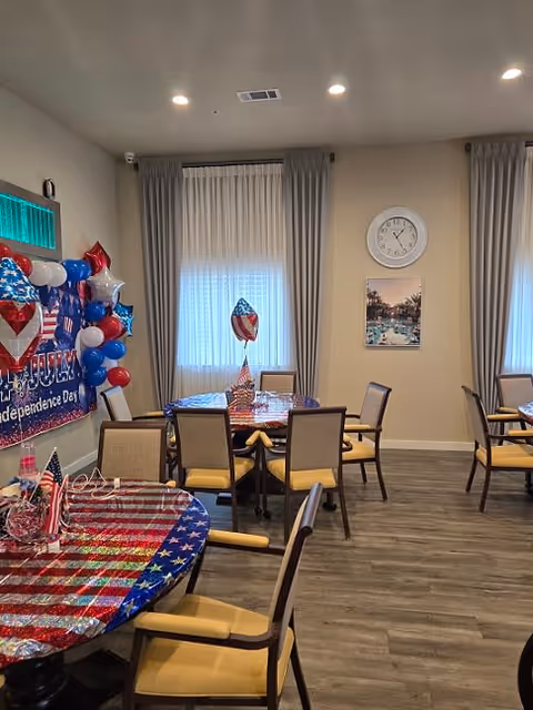 A decorated dining room with round tables covered in red, white, and blue tablecloths featuring stars and stripes. Each table has small American flags and patriotic balloon centerpieces. The room has beige walls, two windows with sheer curtains and gray drapes, a wall clock, and a framed picture. The floor is wood-style, and several chairs with beige cushions surround the tables.