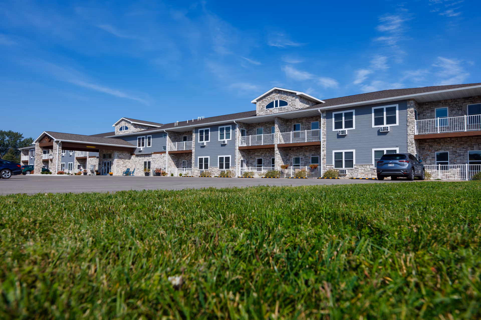 Exterior view of a two-story senior living facility building with stone and gray siding, multiple windows, balconies, and a covered entrance. There are a few cars parked in front and a green lawn in the foreground under a clear blue sky.