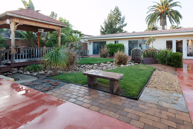 Outdoor garden area at Villa Las Palmas Healthcare Center featuring a wooden gazebo, a stone bench, green grass, various plants, a small wooden bridge, and a building with large windows in the background.