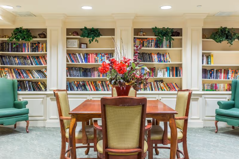 A cozy reading or sitting area with a wooden table surrounded by four chairs. A vase with red and purple flowers is placed on the table. Behind the table, there are built-in bookshelves filled with books and some decorative plants on top. Two green upholstered armchairs are positioned on either side of the bookshelves. The room has a calm and inviting atmosphere.