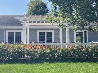 Exterior view of a single-story residential building with light gray siding, white trim, and a pergola over the front porch. There are two windows and a door visible, with a well-maintained green lawn and a row of flowering bushes in front. A large tree provides shade over part of the building.