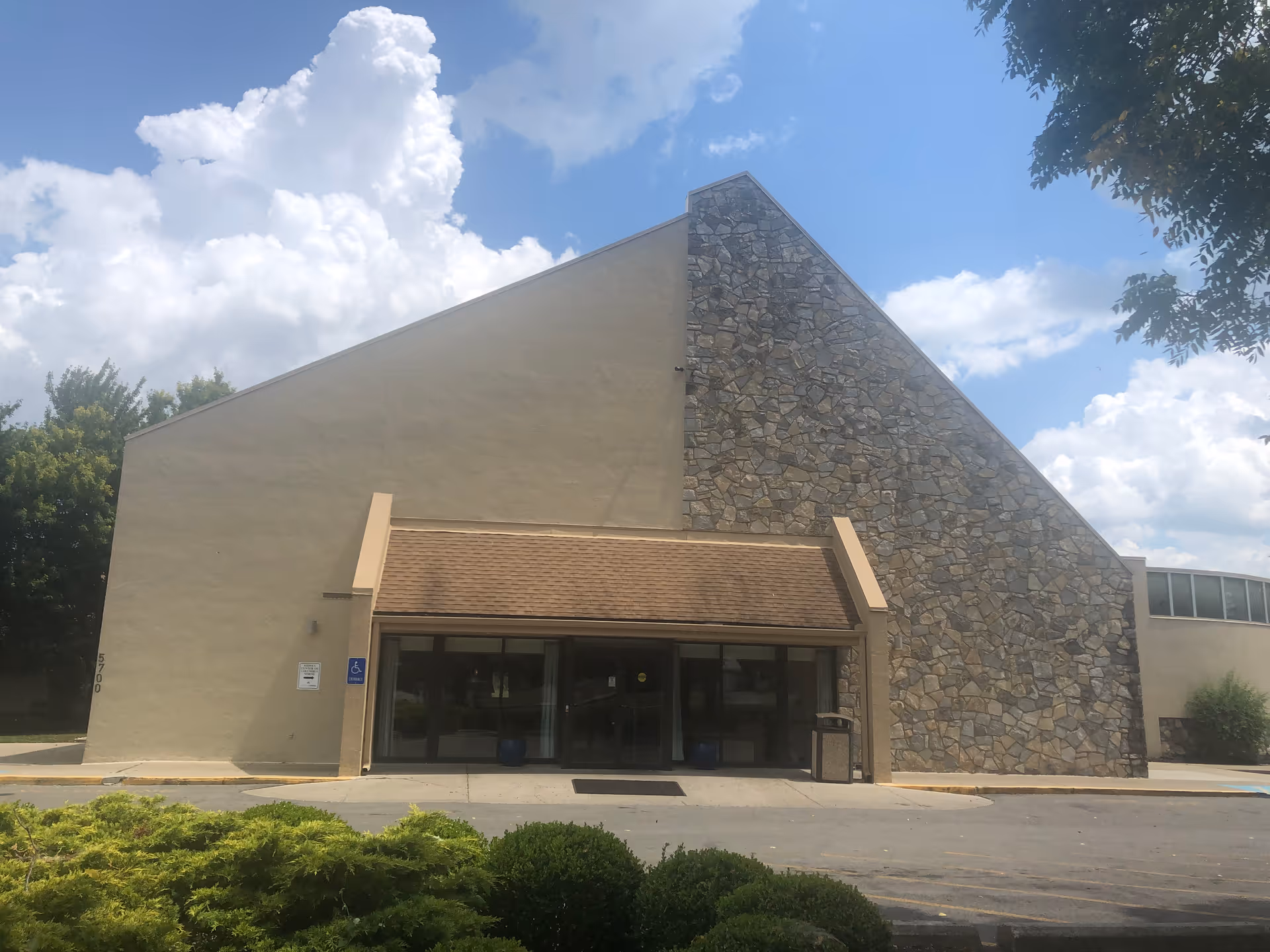 Exterior view of a building with a large triangular stone facade on the right side and a beige wall on the left. The entrance has a small brown awning above glass doors. There are bushes and greenery in the foreground and a partly cloudy sky in the background.