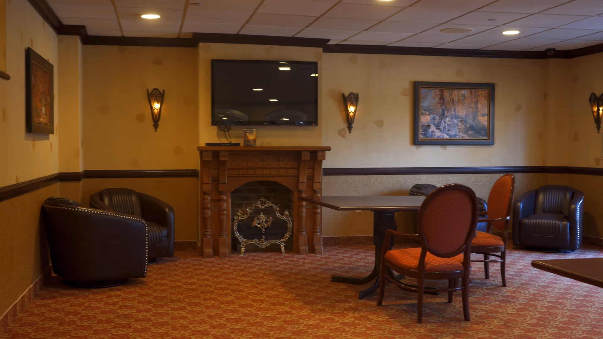 A cozy common area with a patterned red carpet, beige walls, and dark wood trim. The room features a wooden fireplace with a decorative screen, a flat-screen TV mounted above it, two wall sconces with warm lighting, a framed painting on the wall, two dark leather armchairs in the corners, and a table with four red cushioned chairs.