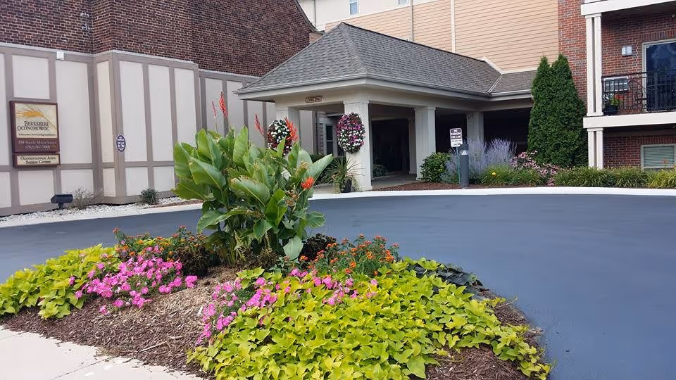 Exterior view of the Berkshire-Oconomowoc senior living facility entrance with a circular driveway and landscaped flower bed featuring green and pink plants. The building has brick and beige paneling with a covered entrance and hanging flower baskets.