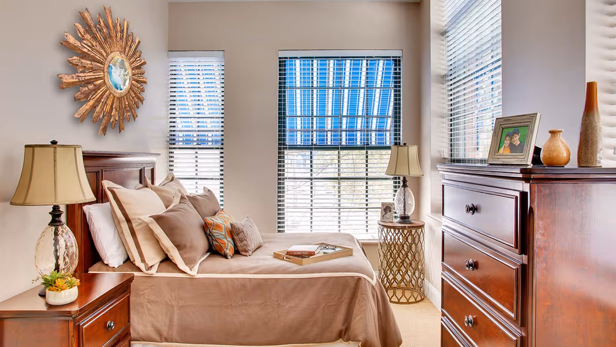 A cozy bedroom with a neatly made bed featuring multiple pillows in neutral tones. There are two wooden nightstands with lamps on either side of the bed. A decorative sunburst mirror hangs on the wall above the headboard. The room has three windows with white blinds and a blue striped valance, allowing natural light to fill the space. A wooden dresser with framed photos and decorative vases is positioned near the windows.