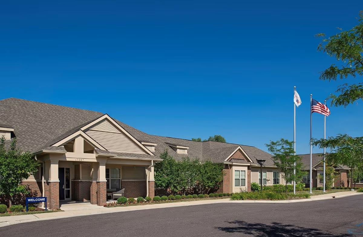 Exterior view of a single-story senior living facility building with a welcoming entrance, well-maintained landscaping, and two flagpoles displaying the American flag and another flag under a clear blue sky.