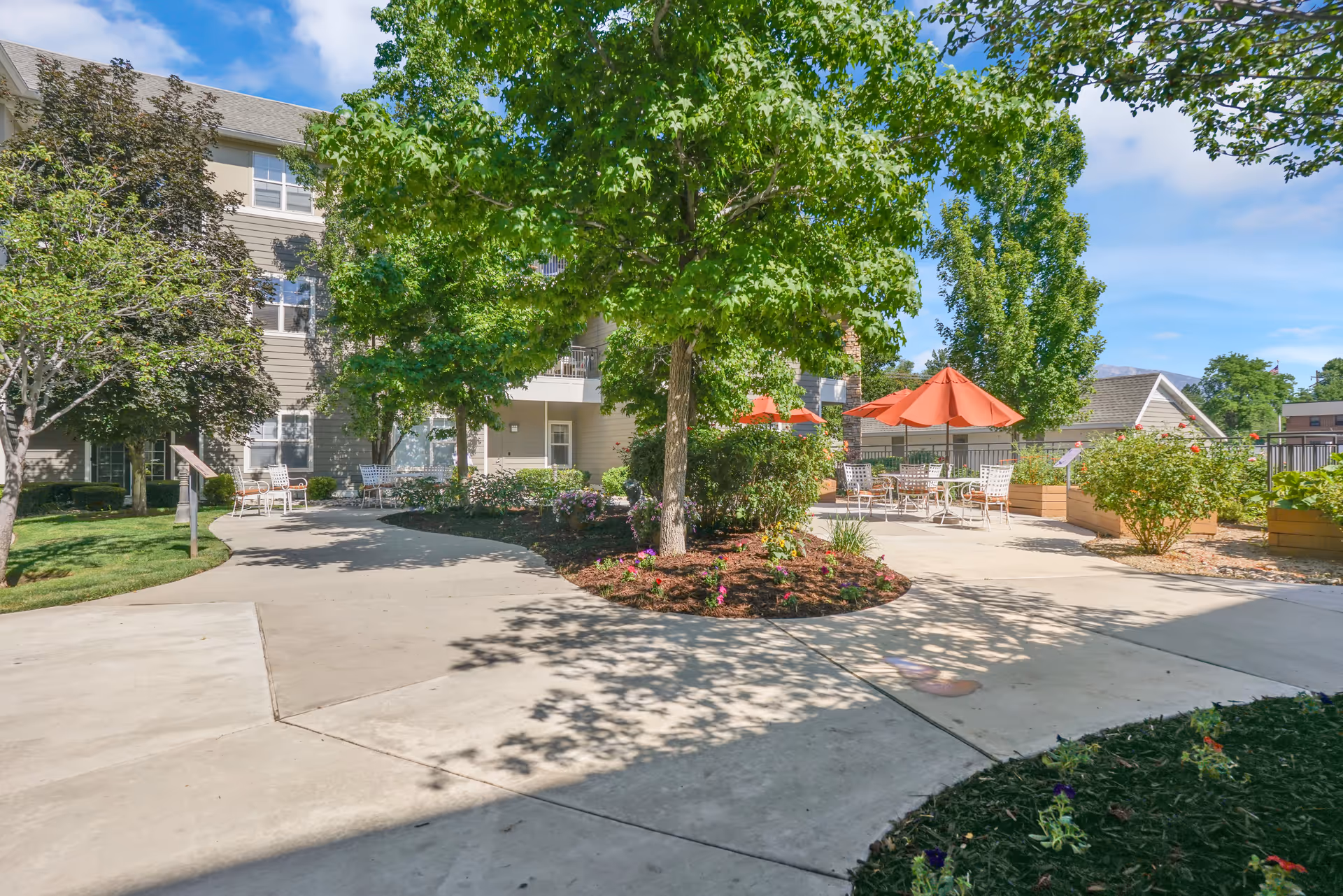 Outdoor courtyard area at The Wellington featuring a paved walkway, green trees, bushes, and flower beds. There are several white metal tables and chairs with red umbrellas providing shade. The building exterior is visible in the background under a blue sky with some clouds.