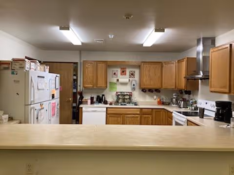 A kitchen with wooden cabinets, a white refrigerator covered with papers and magnets, a white stove with a stainless steel range hood, a coffee maker, and various kitchen items on the countertops. The kitchen is viewed from behind a beige countertop bar.