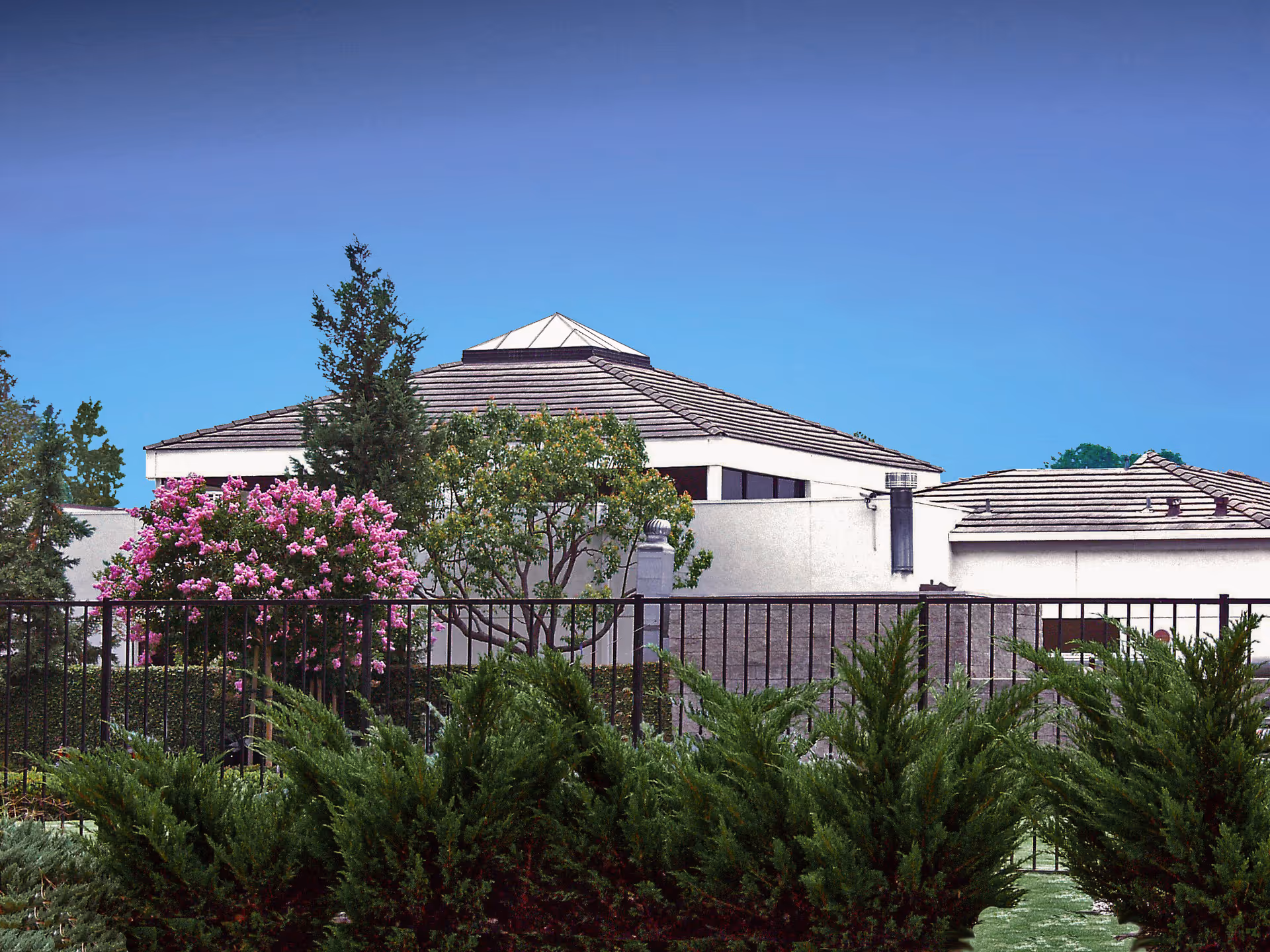 Exterior view of a building with a tiled roof and a small glass pyramid structure on top, surrounded by trees and flowering bushes, with a black metal fence in the foreground under a clear blue sky.