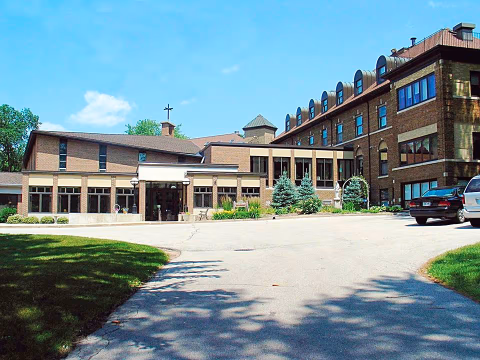 Exterior view of McCormick Assisted Living facility showing a large brick building with multiple windows, a driveway, parked cars, and some greenery including trees and bushes under a clear blue sky.