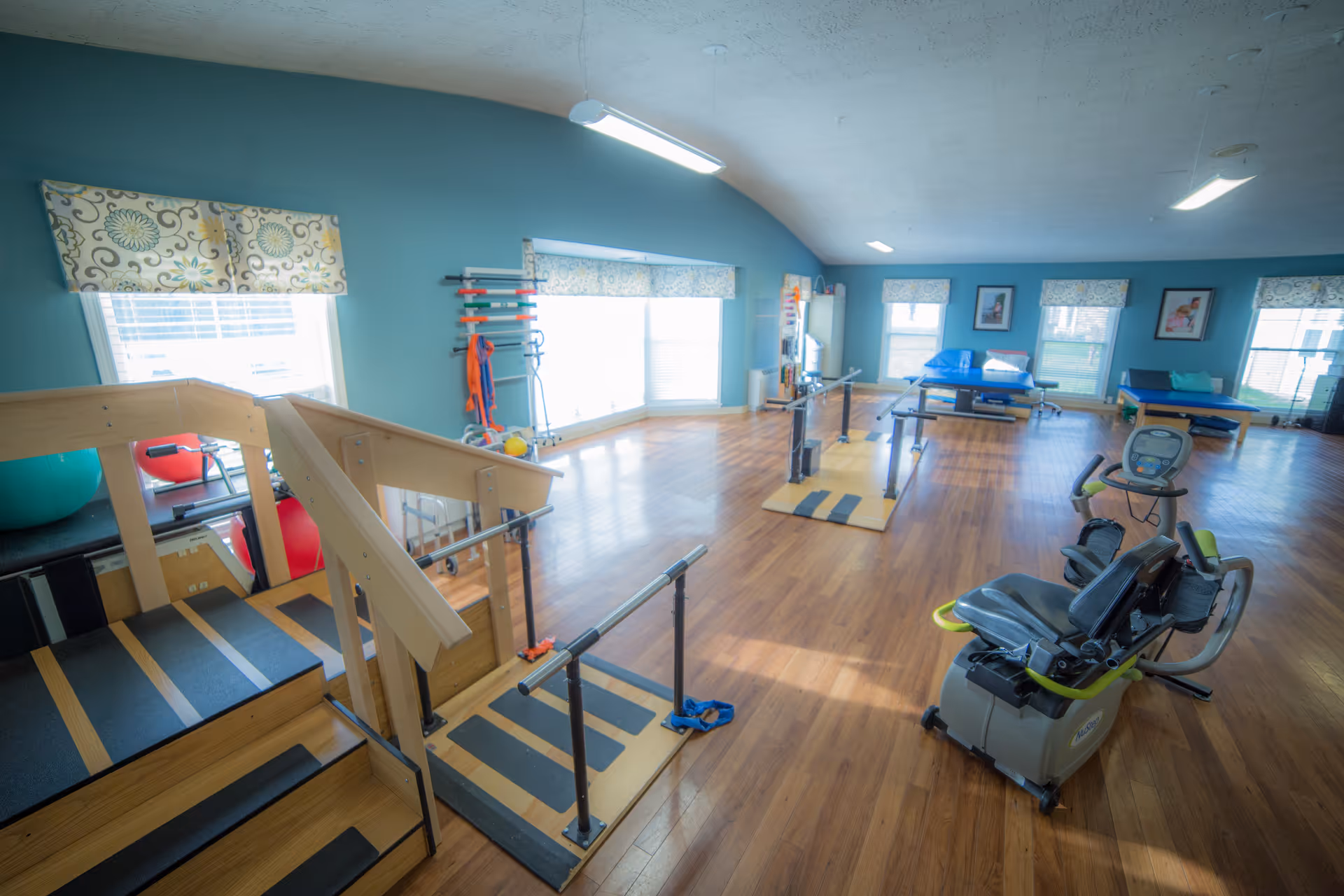 A spacious rehabilitation room with wooden flooring and blue walls. The room contains exercise equipment including parallel bars, a recumbent exercise bike, therapy tables, and a wooden staircase for physical therapy. Large windows with patterned valances allow natural light to fill the room.
