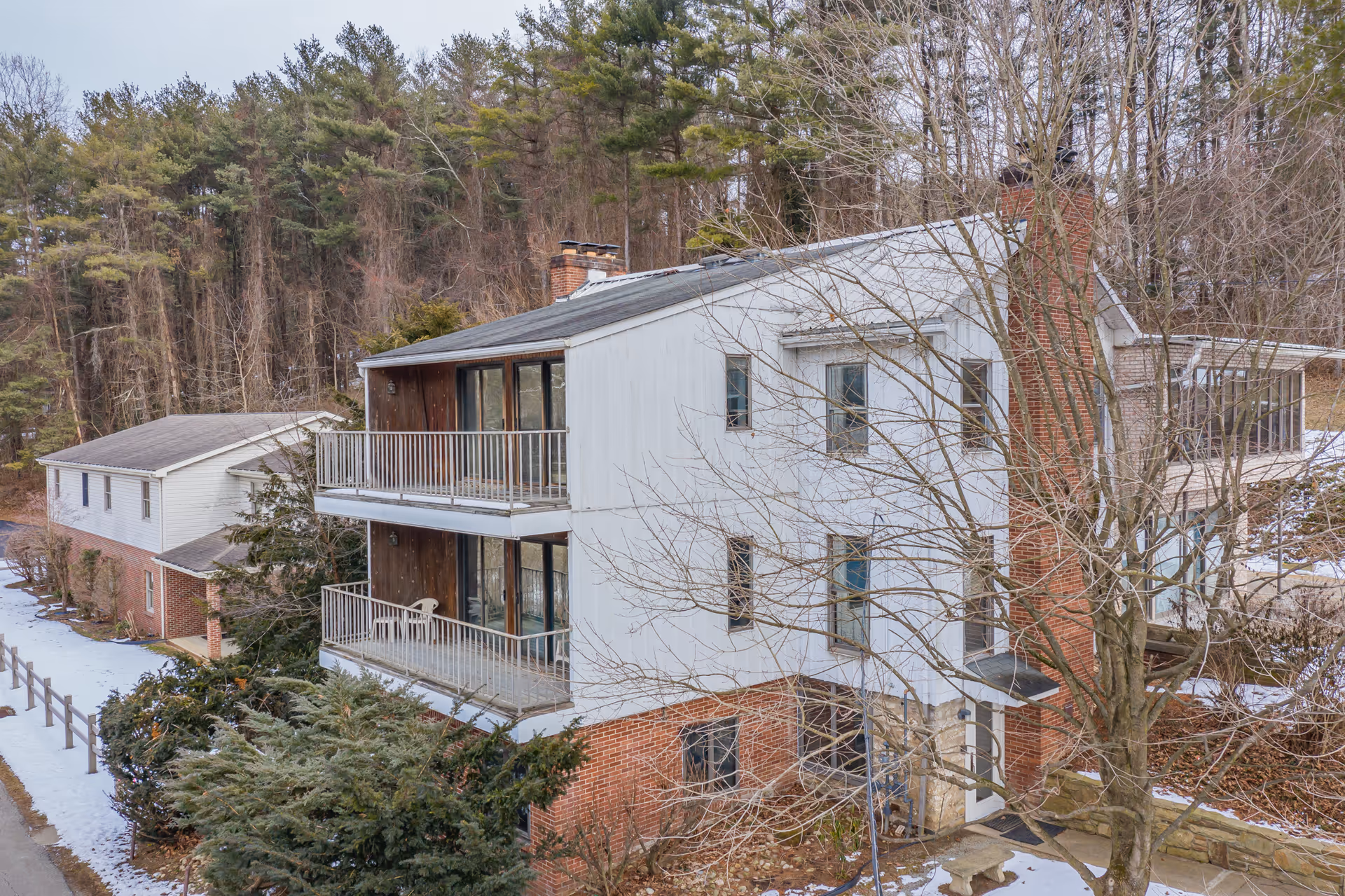 Exterior view of a multi-story residential building with balconies, surrounded by trees and light snow on the ground. The building has a brick lower level and white upper levels with large windows and a chimney. Another building is visible in the background.