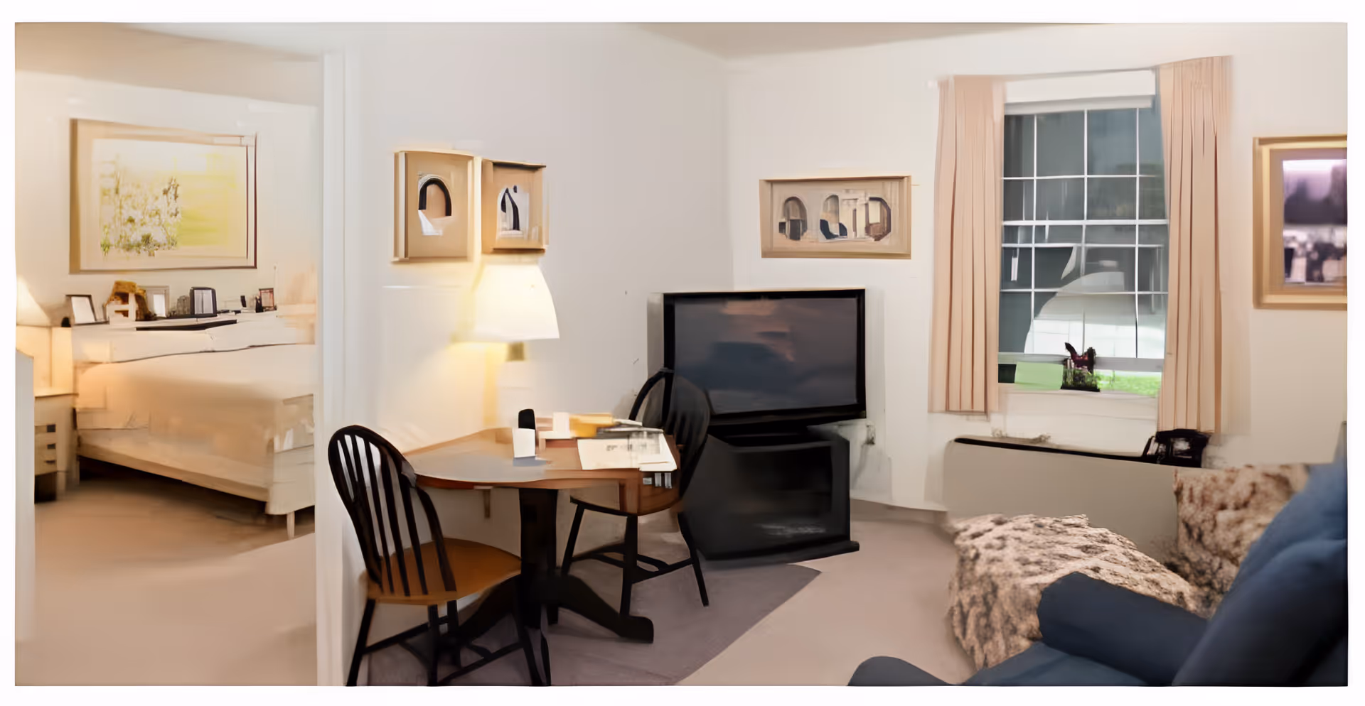 A cozy living room area with a small wooden table and two chairs, a flat-screen TV on a stand, and a window with beige curtains letting in natural light. To the left, a bedroom with a bed and nightstand is visible through an open doorway. The room has light-colored walls and carpeted floors, with framed artwork on the walls.