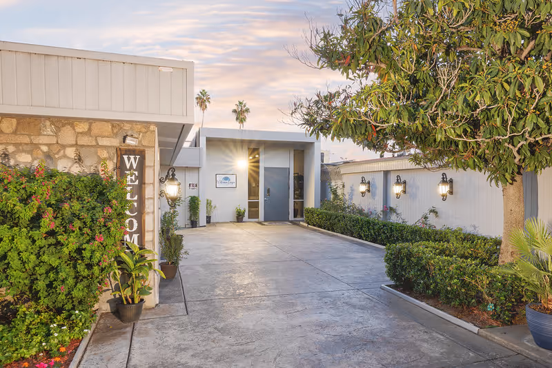Entrance to a building at Waterman Canyon Post Acute with a concrete pathway leading to a door. The area is landscaped with green bushes, potted plants, and a tree. A vertical wooden sign on the left reads 'WELCOME'. The building exterior is light-colored with wall-mounted lantern-style lights and a partly cloudy sky at sunset.