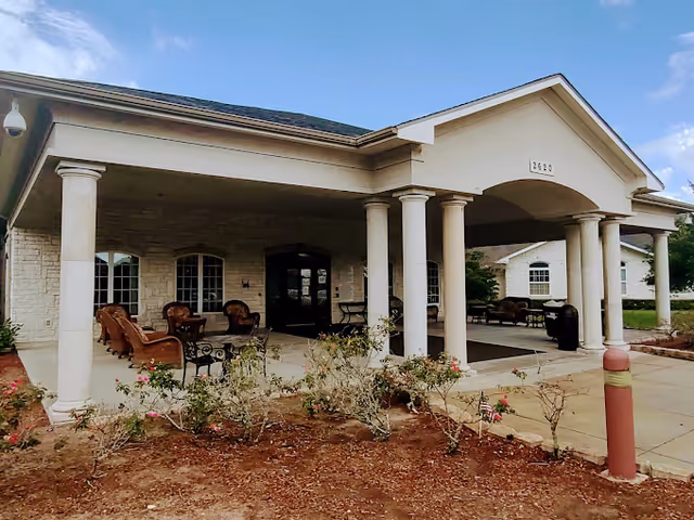Front entrance of The Heights of League City facility featuring a covered porch with white columns, outdoor seating with chairs and tables, and a garden area with small plants and flowers in front. The building has a light-colored brick exterior and a peaked roof under a partly cloudy sky.