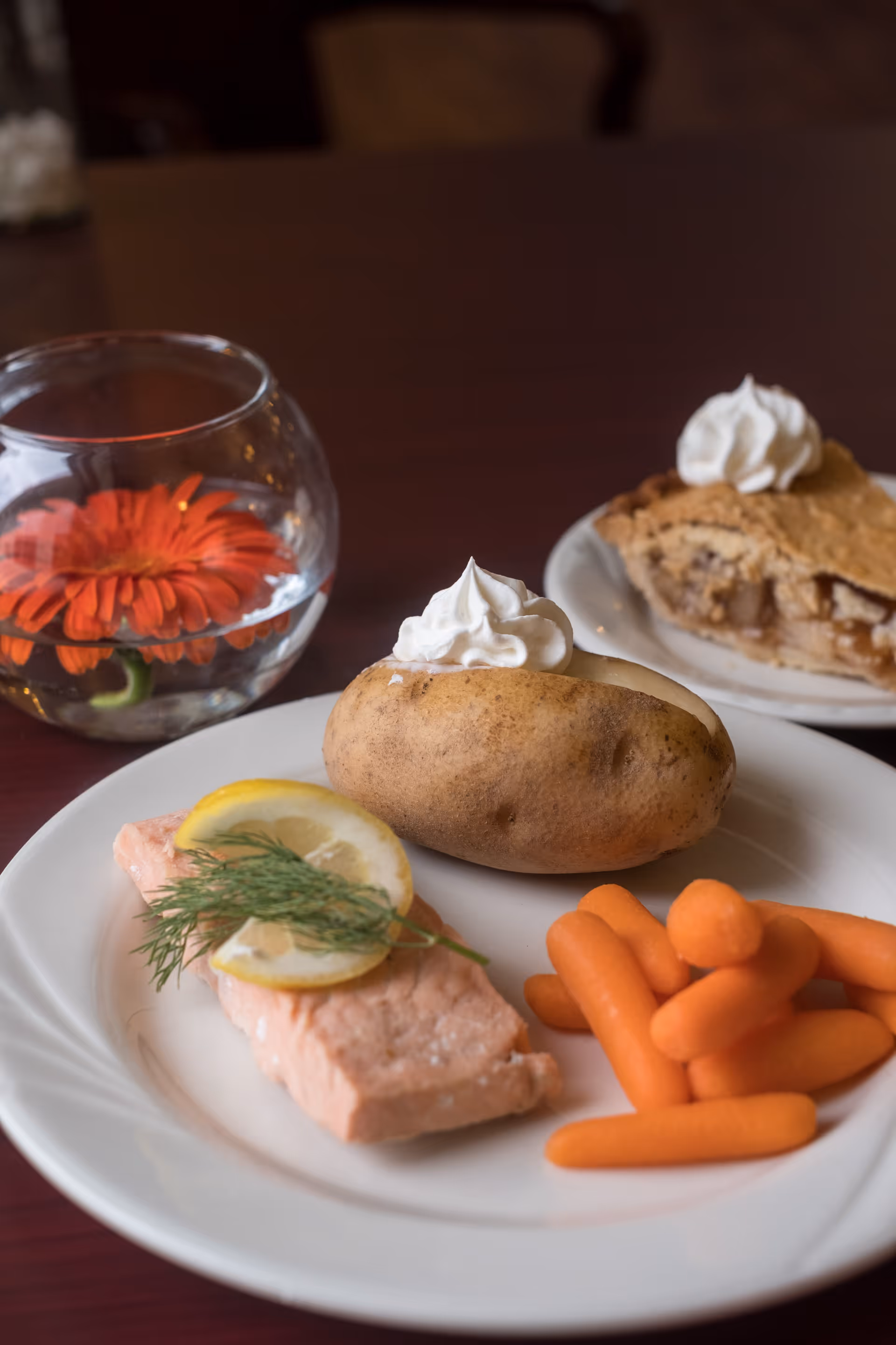 A plate with a baked potato topped with sour cream, a piece of cooked salmon garnished with lemon slices and dill, and a serving of baby carrots. In the background, there is a slice of apple pie with whipped cream on a separate plate and a small round glass vase with an orange flower and water.