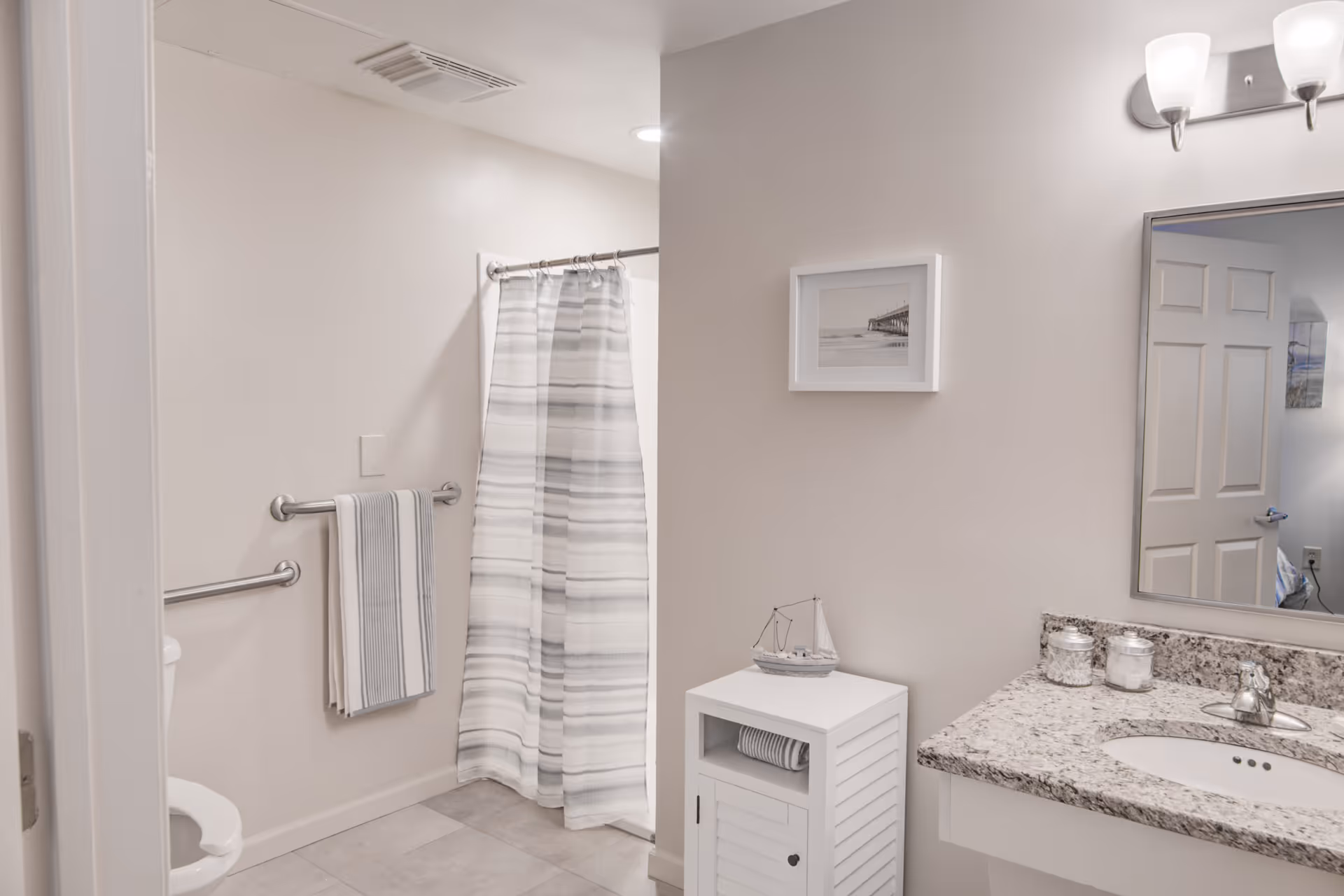 A clean and bright bathroom featuring a granite countertop with a sink, a large mirror, a small white cabinet with a decorative sailboat on top, a towel rack with a striped towel, a toilet, and a shower with a striped curtain.