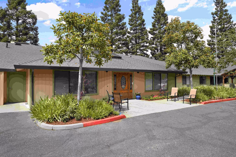 Exterior view of Almond Grove Assisted Living facility showing a single-story building with a gray roof, beige and green walls, and a wooden front door with decorative glass. There are two small trees and landscaped bushes in front, along with outdoor chairs and a paved driveway under a partly cloudy sky.