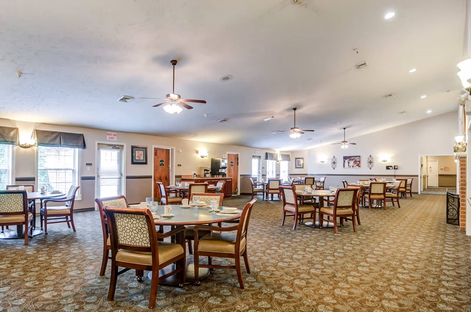 Spacious dining room with multiple round tables and chairs arranged neatly. The room has carpeted flooring, ceiling fans with lights, and large windows letting in natural light. Walls are decorated with framed pictures and sconces. There is a door marked 'Not an Exit' and a small counter area in the background.