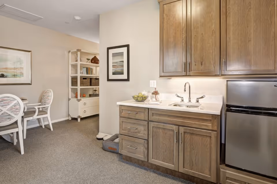 Interior view of a senior living facility showing a kitchenette area with wooden cabinets, a small stainless steel refrigerator, and a sink with a faucet. To the left, there is a dining area with a round table and two chairs with patterned cushions. A white shelving unit with decorative items and books is visible in the background, along with framed artwork on the walls.