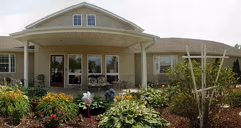 Front exterior view of a single-story building with a covered porch, several windows, and a door. The porch has chairs and a bench, and there are various plants and flowers in a garden bed in front of the building.