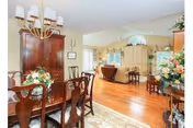 A spacious dining room with a wooden dining table and chairs, a chandelier overhead, and floral arrangements on the table and side furniture. The room opens into a living area with hardwood floors, a fireplace, and additional seating. The walls are painted in light colors, and there are large windows allowing natural light to fill the space.
