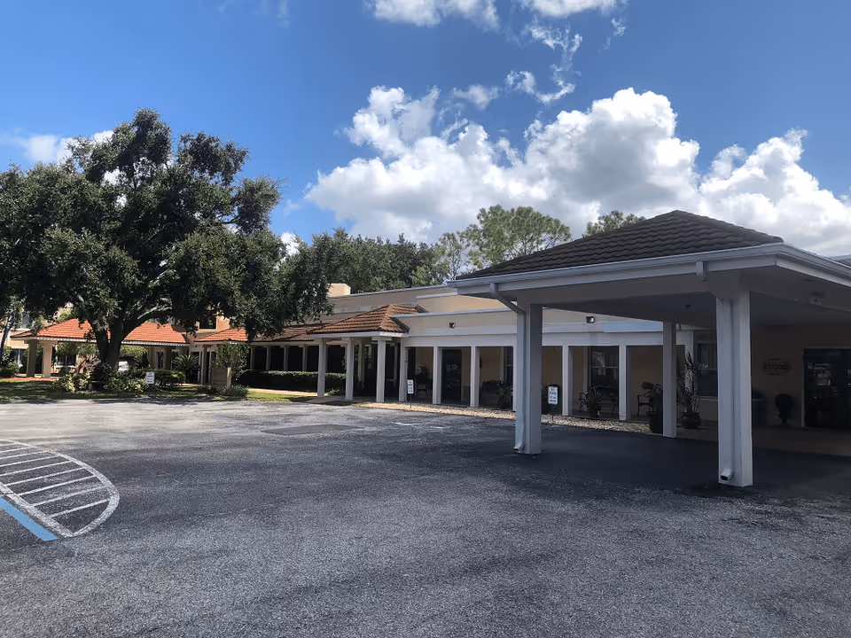 Exterior view of Lake Mariam Health and Rehabilitation Center showing a single-story building with a covered entrance, large trees, and a parking area under a partly cloudy blue sky.