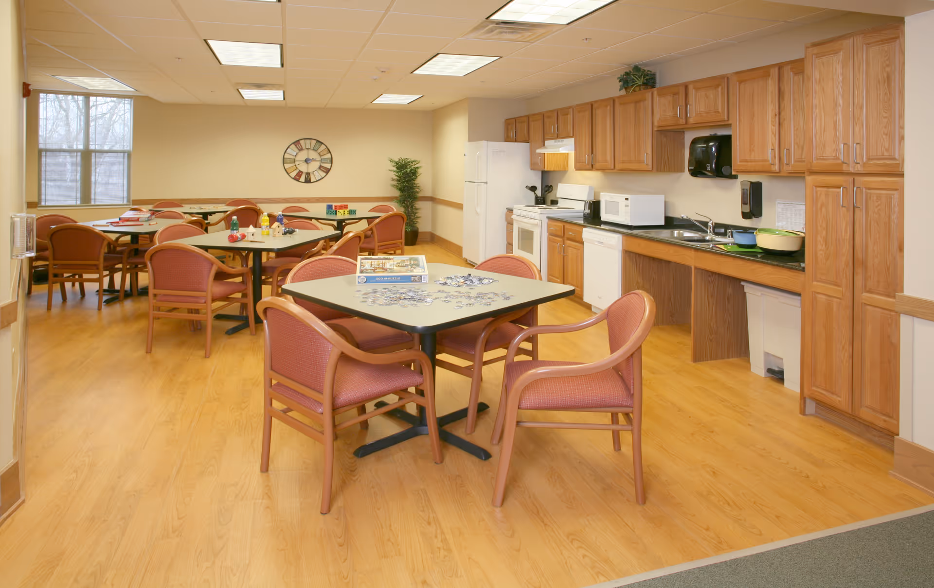 A spacious room with multiple tables and chairs arranged for dining or activities. The room features a kitchenette area with wooden cabinets, a refrigerator, microwave, stove, and sink. There is a large clock on the wall and a window letting in natural light. The floor is wood laminate, and the ceiling has recessed lighting.