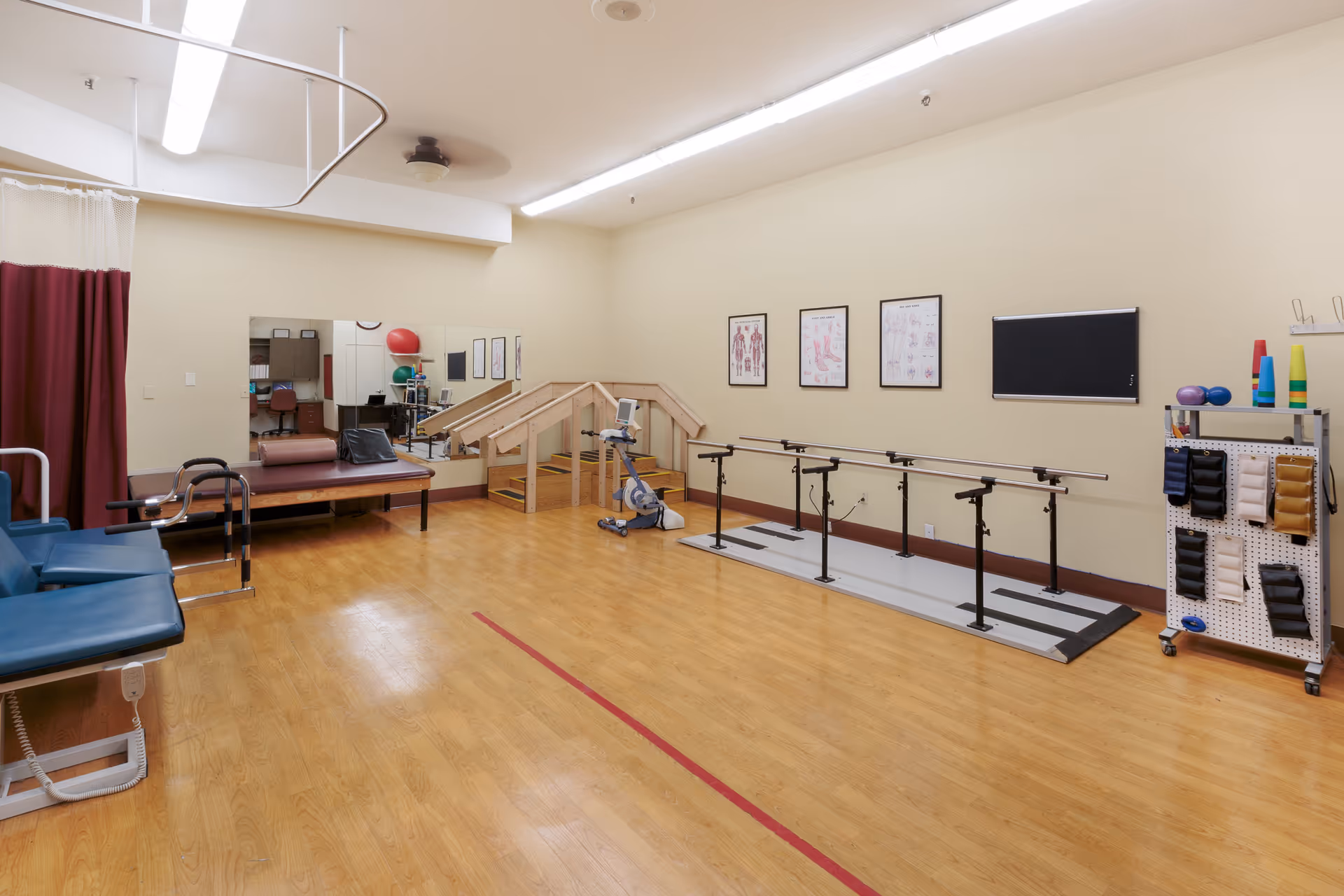 A physical therapy room with wooden flooring, parallel bars for walking exercises, a stationary exercise bike, a set of stairs with handrails, a padded examination table, and a rack holding various therapy equipment such as weights and cones. The walls have anatomical charts and a mirror reflecting part of the room.