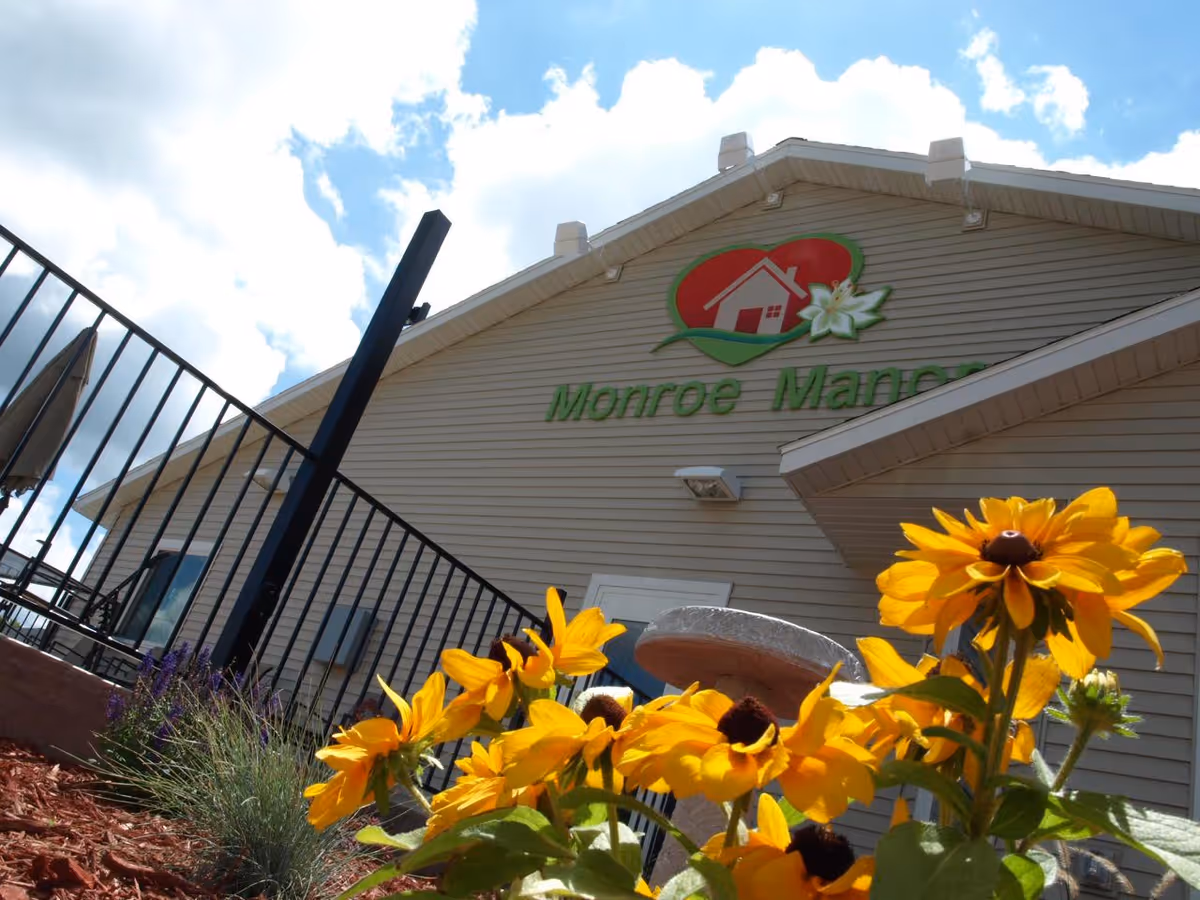 View of the exterior of Monroe Manor building with a clear blue sky and white clouds above. Bright yellow flowers are in the foreground near a black metal fence and a birdbath or similar structure.