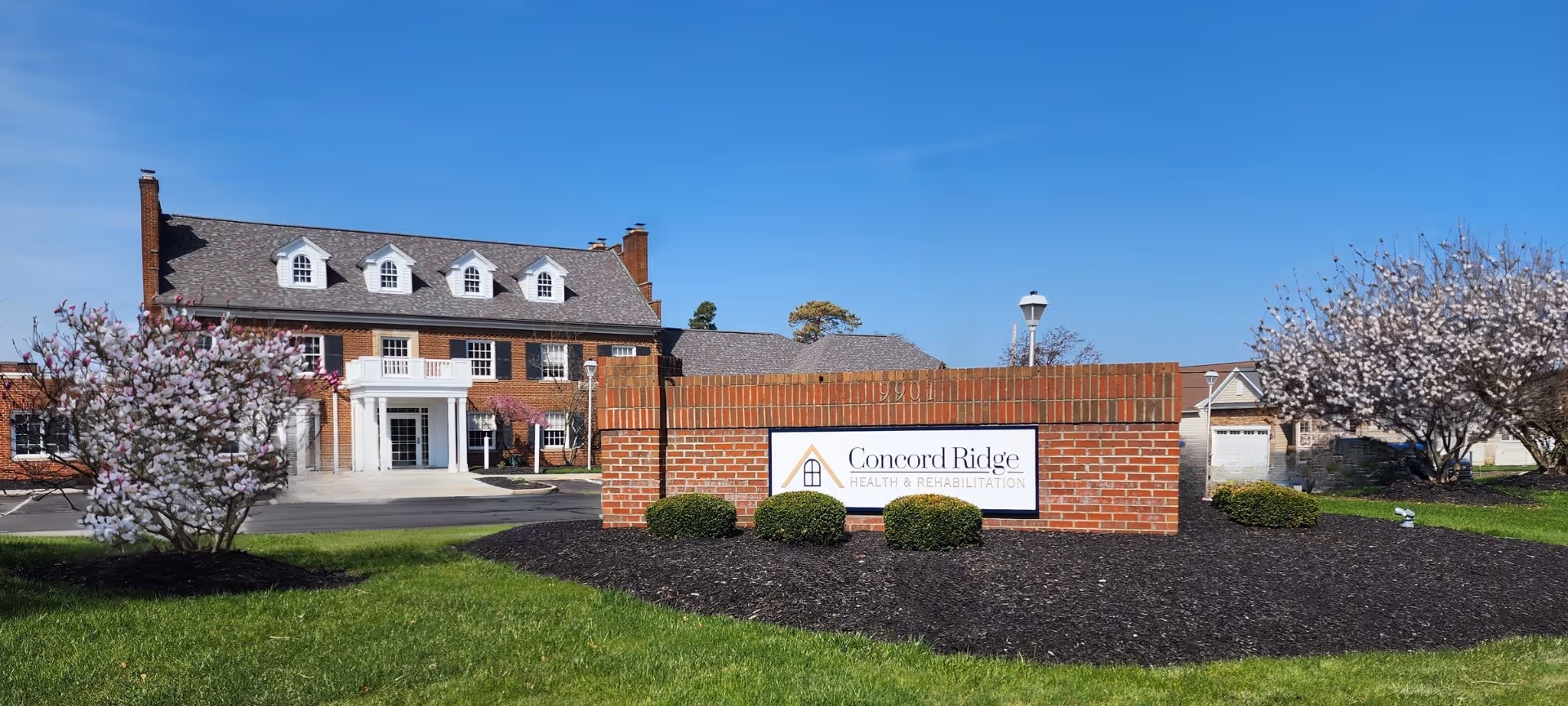 Front exterior view of Concord Ridge Health & Rehabilitation building with a brick sign in front, surrounded by green grass and blooming trees under a clear blue sky.