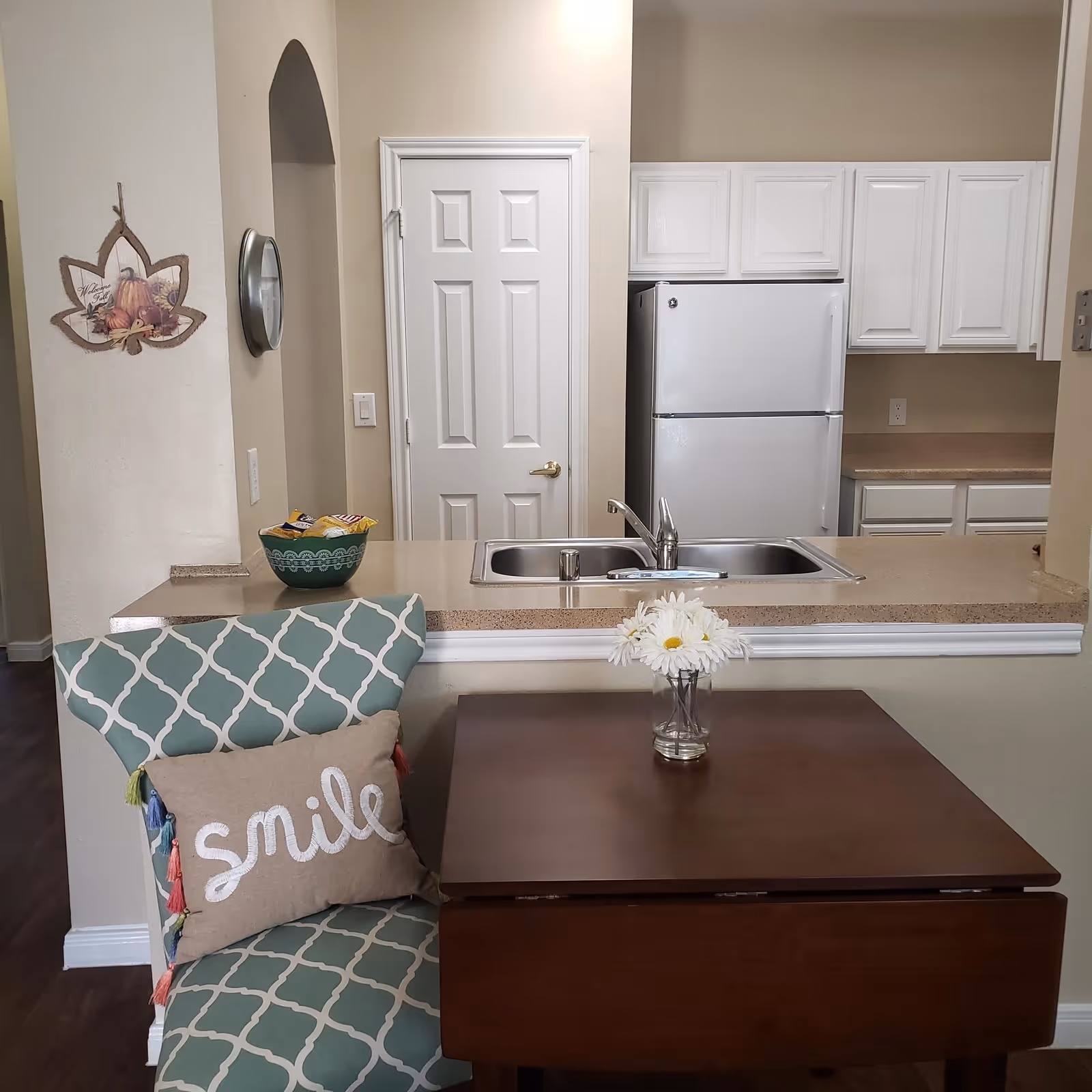 A small kitchen area with white cabinets and a white refrigerator. A double sink is set into a beige countertop that extends into a breakfast bar. In front of the bar is a wooden table with a glass vase holding white daisies. A cushioned chair with a green and white geometric pattern and a pillow that says 'smile' is positioned next to the table. On the wall to the left, there is a decorative hanging shaped like a leaf with a fall theme and a round wall clock.