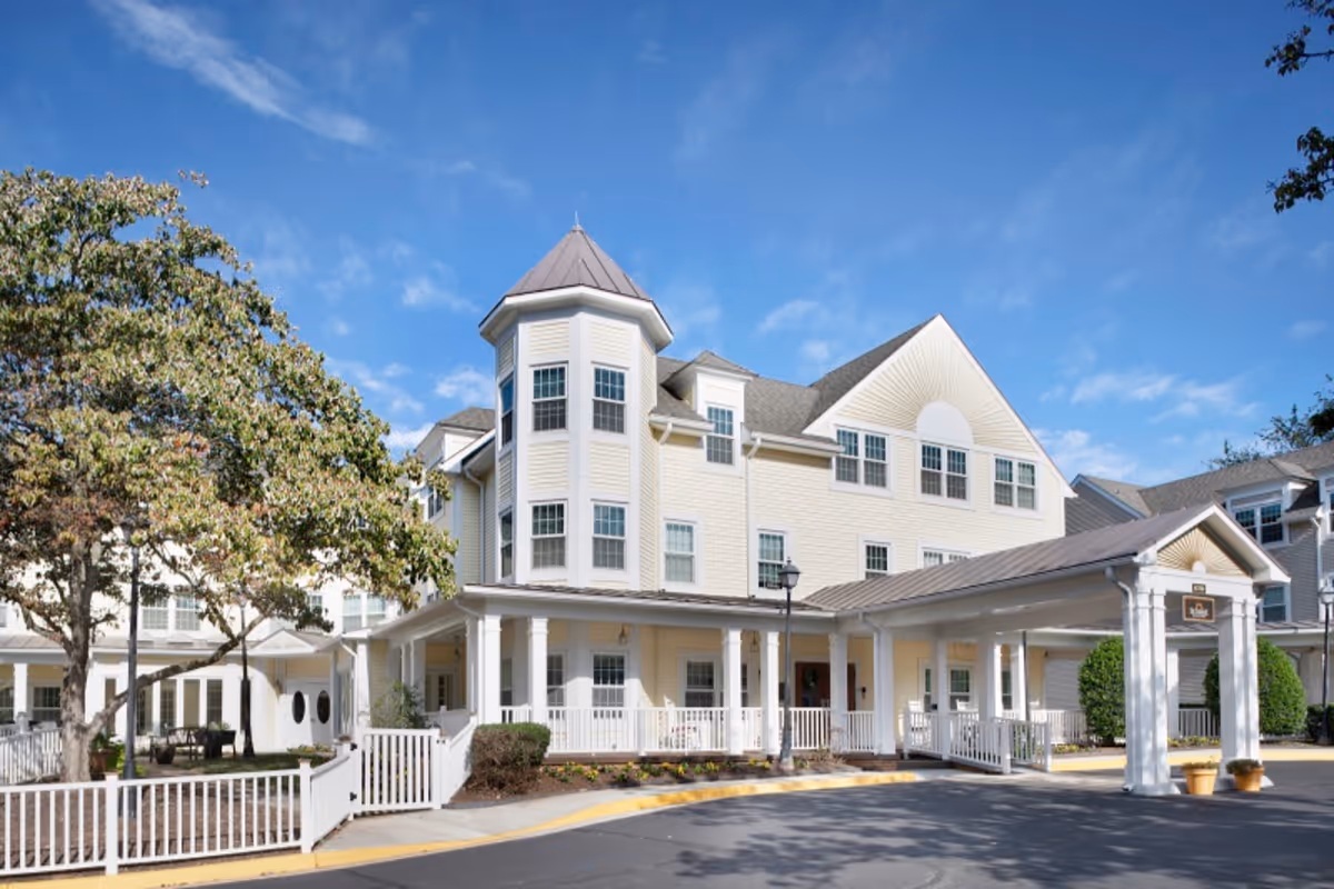 Exterior view of a large, multi-story senior living facility with light yellow siding, white trim, and a covered entrance. There is a tree and white fencing in the foreground under a clear blue sky.