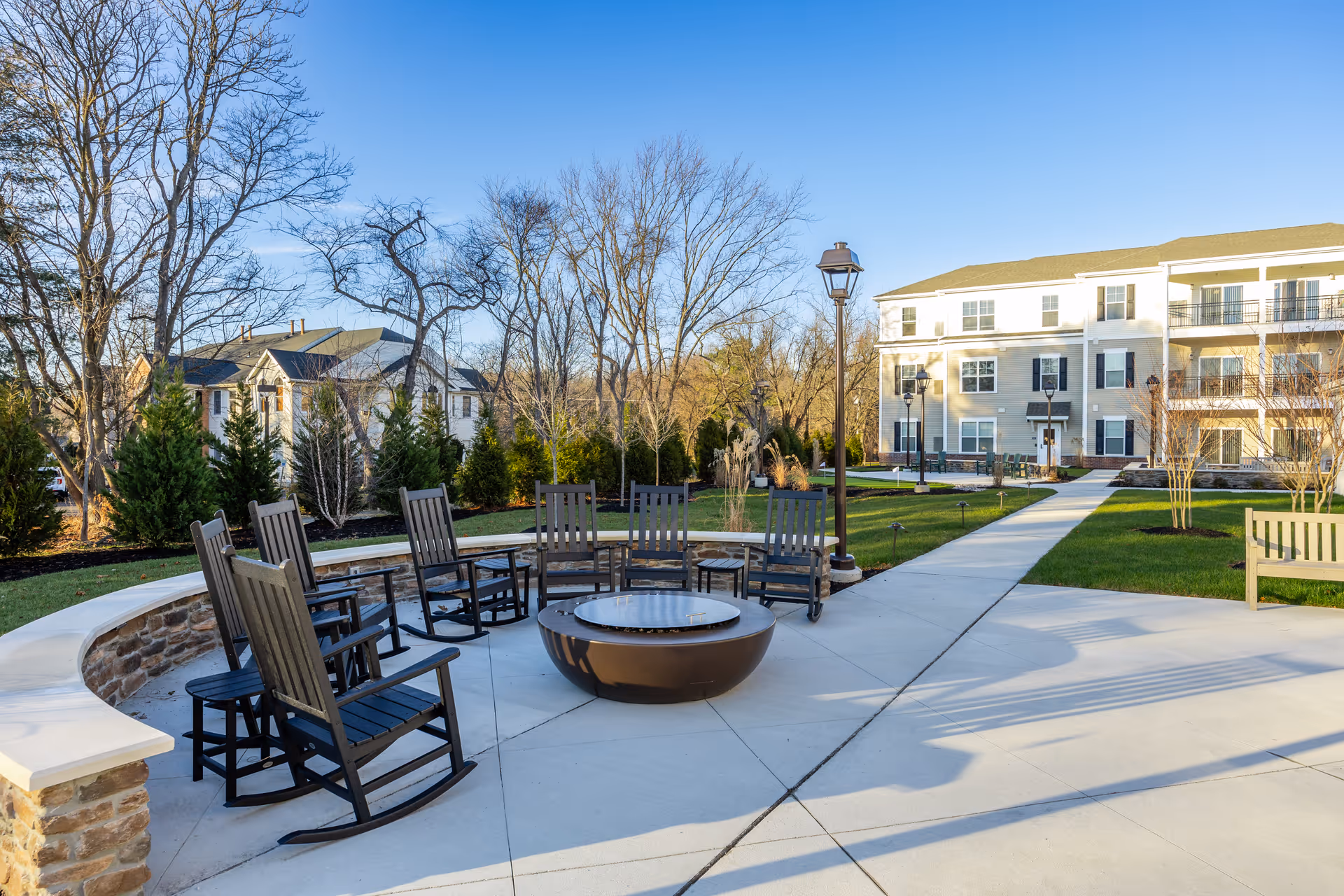 Outdoor seating area with black rocking chairs arranged in a semicircle around a fire pit on a concrete patio. There is a stone wall bench curving around part of the seating area. In the background, there are trees without leaves, a grassy lawn, a paved walkway, and a multi-story residential building with balconies under a clear blue sky.