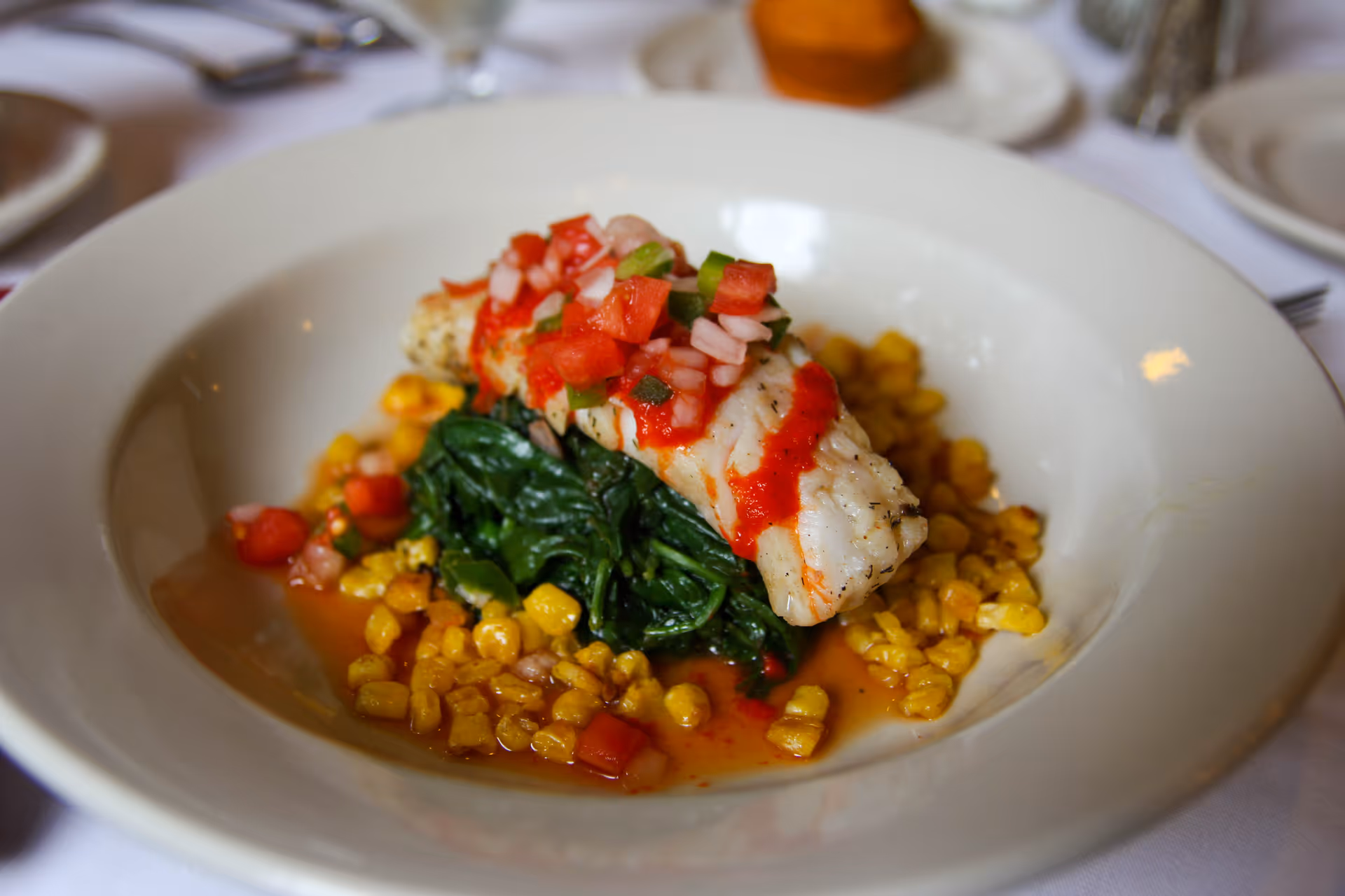 A plated dish featuring a piece of grilled white fish topped with diced tomatoes and onions, served on a bed of sautéed spinach and surrounded by cooked corn kernels in a shallow white bowl.