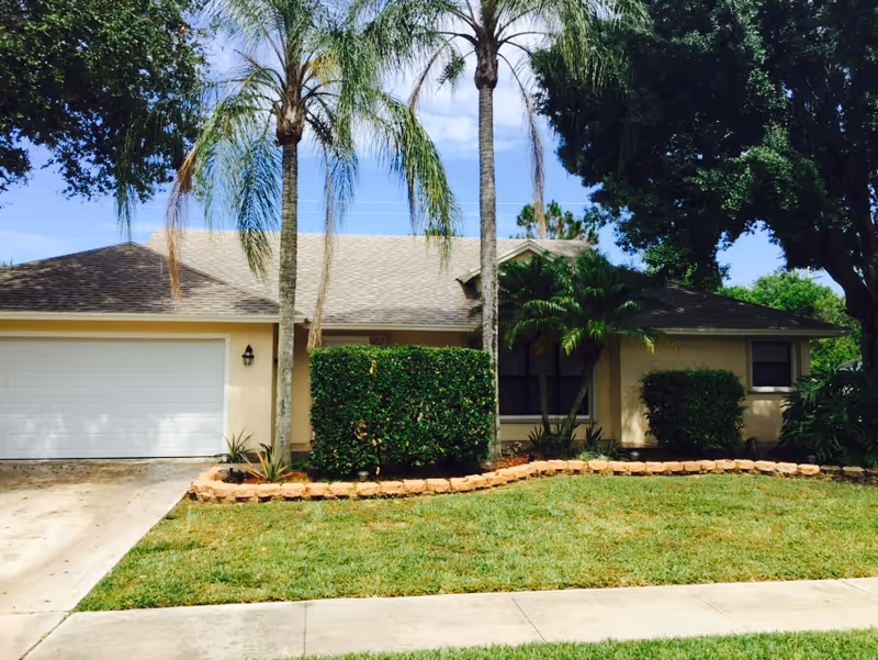 Single-story house with a beige exterior, a two-car garage, and a well-maintained front yard featuring palm trees, bushes, and a stone border along the garden bed.