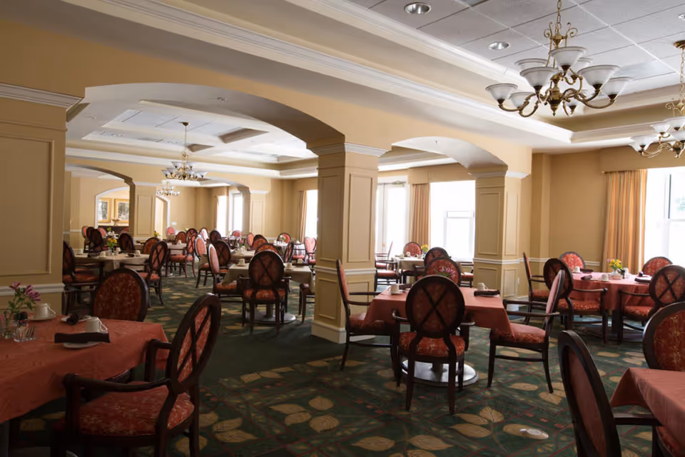 A spacious dining room with multiple round tables covered with red tablecloths and set with cups and napkins. The room features elegant chandeliers, large windows with beige curtains, and green carpet with a leaf pattern. The walls are painted in a warm beige tone with white trim and columns dividing the space.