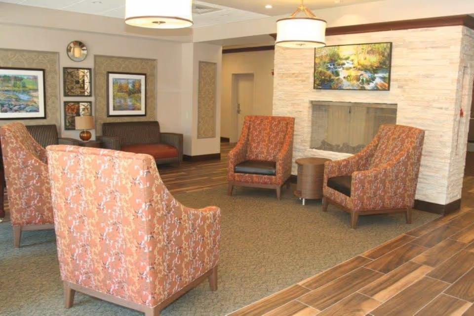 A cozy senior living facility lounge area with four patterned armchairs arranged around a small round wooden table in front of a modern stone fireplace. The room features wood-look tile flooring, a carpeted seating area, framed landscape paintings on the walls, and soft overhead lighting fixtures.
