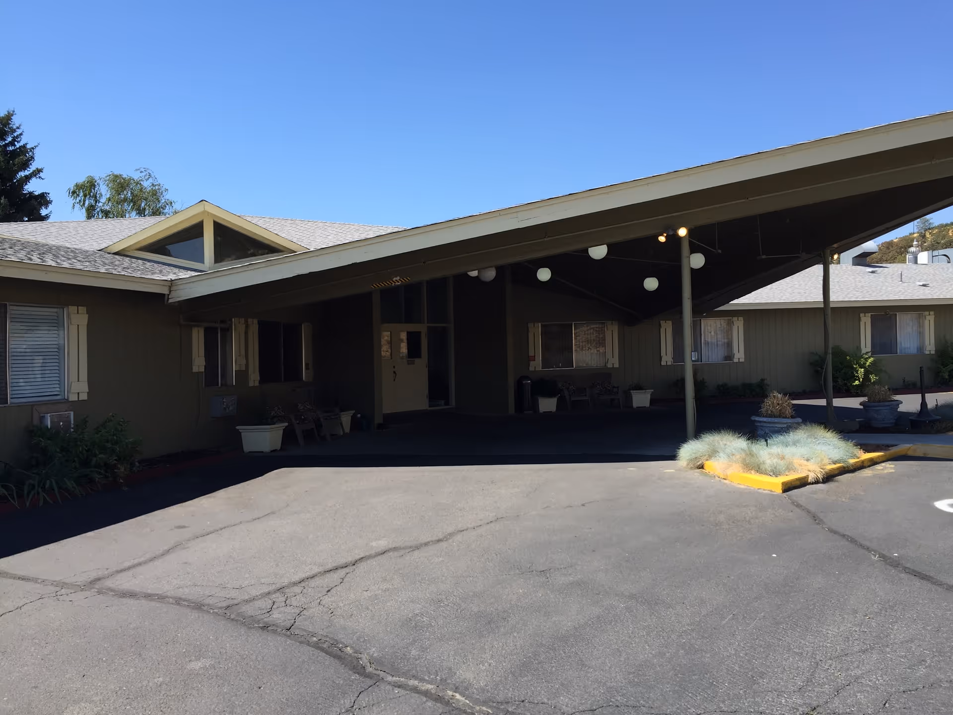 Exterior view of The Dalles Health and Rehabilitation facility showing the entrance with a large covered drop-off area, beige building walls, several windows with shutters, potted plants, and a clear blue sky.